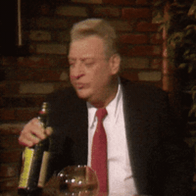 A Man In A Suit And Tie Is Sitting At A Table Holding A Bottle Of Wine GIF