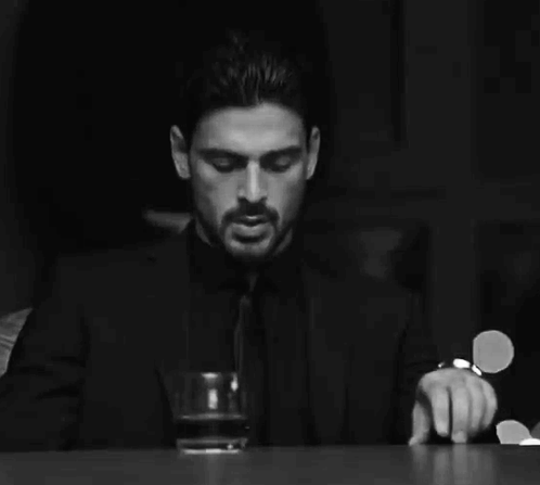 A Man In A Suit And Tie Is Sitting At A Table Holding A Glass Of Whiskey . GIF