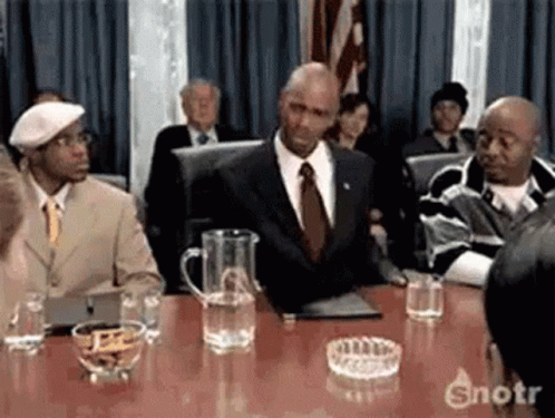 A Man In A Suit And Tie Is Sitting At A Table In Front Of A Crowd . GIF