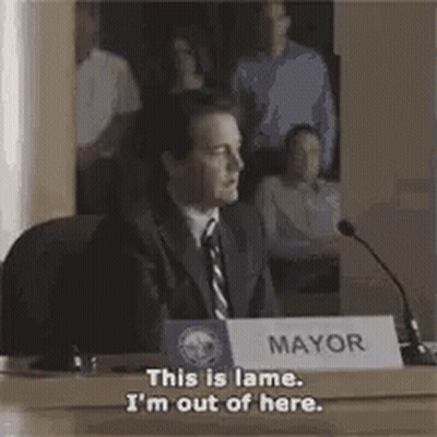 A Man In A Suit And Tie Is Sitting At A Table In Front Of A Microphone And A Sign That Says Mayor . GIF