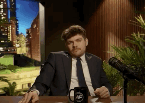 A Man In A Suit And Tie Is Sitting At A Table In Front Of A Microphone With A Mug On It . GIF