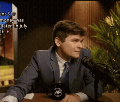 A Man In A Suit And Tie Is Sitting At A Table In Front Of A Microphone With A Mug That Says Af On It GIF