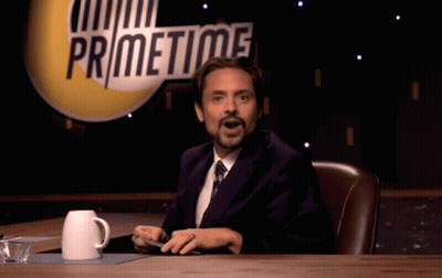 A Man In A Suit And Tie Is Sitting At A Table In Front Of A Sign That Says Primetime GIF
