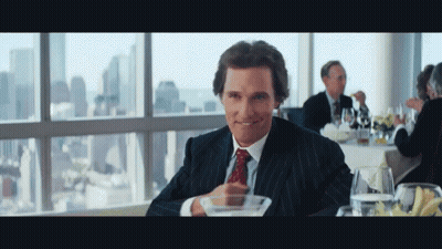 A Man In A Suit And Tie Is Sitting At A Table In Front Of A Window GIF