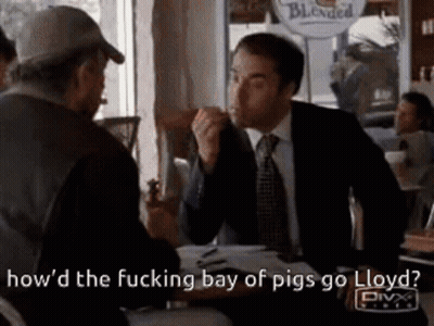 A Man In A Suit And Tie Is Sitting At A Table Talking To Another Man In A Hat . GIF