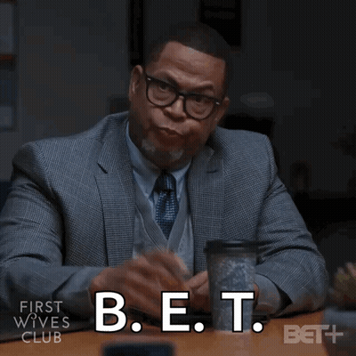 A Man In A Suit And Tie Is Sitting At A Table With A Cup Of Coffee And The Words B.e.t. Below Him GIF