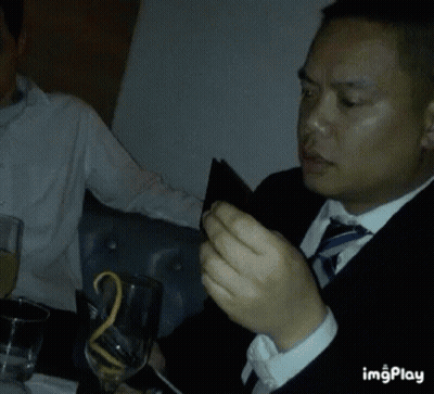 A Man In A Suit And Tie Is Sitting At A Table With A Glass Of Wine And A Wallet . GIF