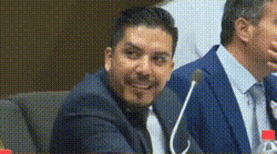 A Man In A Suit And Tie Is Sitting At A Table With A Microphone . GIF