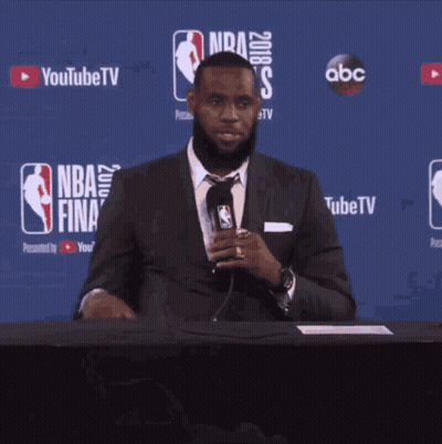 A Man In A Suit And Tie Is Sitting At A Table With A Microphone In Front Of A Nba Finals Sign GIF