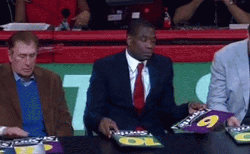 A Man In A Suit And Tie Is Sitting At A Table With A Sign That Says Gatorade On It . GIF