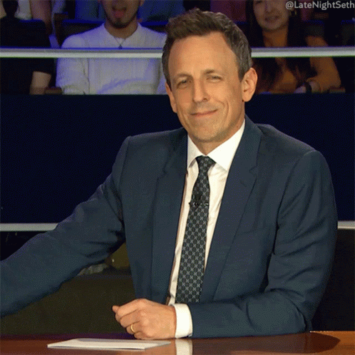 A Man In A Suit And Tie Is Sitting At A Table With His Arms Crossed In Front Of A Crowd GIF