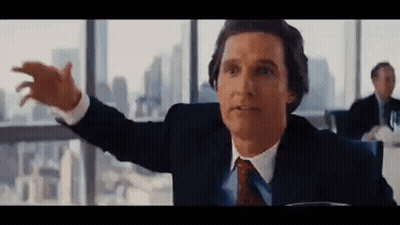 A Man In A Suit And Tie Is Sitting At A Table With His Arms Outstretched In Front Of A Window . GIF