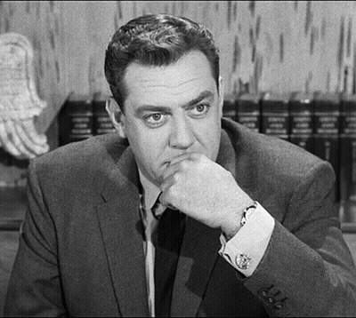 A Man In A Suit And Tie Is Sitting At A Table With His Hand On His Chin And Looking At The Camera . GIF