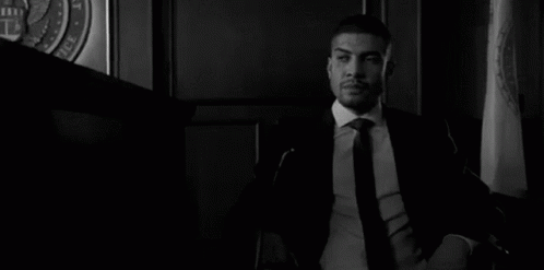 A Man In A Suit And Tie Is Sitting At A Table With His Hands Folded In A Courtroom . GIF