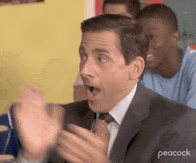 A Man In A Suit And Tie Is Sitting At A Table With His Hands In The Air And His Mouth Open . GIF