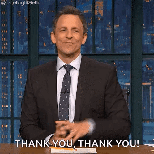 A Man In A Suit And Tie Is Sitting At A Table With His Hands In The Air And Saying Thank You . GIF