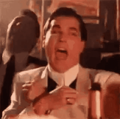 A Man In A Suit And Tie Is Sitting At A Table With His Mouth Open And Holding A Glass Of Wine . GIF