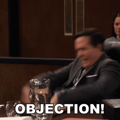 A Man In A Suit And Tie Is Sitting At A Table With Objection Written On It GIF