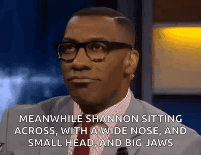 A Man In A Suit And Tie Is Sitting Down With A Wide Nose And A Small Head And Big Jaws . GIF