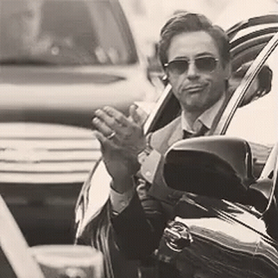 A Man In A Suit And Tie Is Sitting In A Car Waving His Hand . GIF