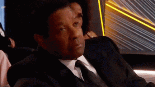 A Man In A Suit And Tie Is Sitting In A Chair And Looking At Something . GIF
