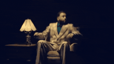 A Man In A Suit And Tie Is Sitting In A Chair In Front Of A Lamp GIF