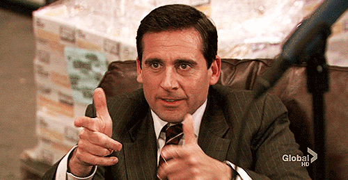 A Man In A Suit And Tie Is Sitting In A Chair Pointing At The Camera . GIF