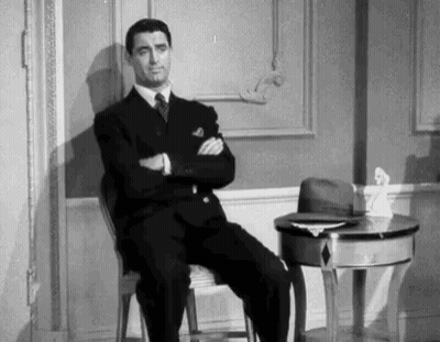 A Man In A Suit And Tie Is Sitting In A Chair With His Arms Crossed In A Room . GIF