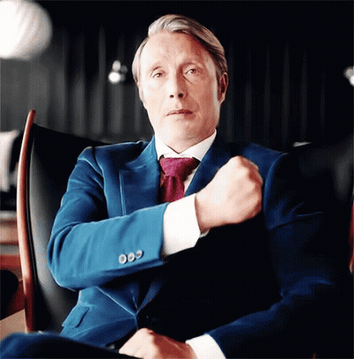 A Man In A Suit And Tie Is Sitting In A Chair With His Fist In His Pocket . GIF