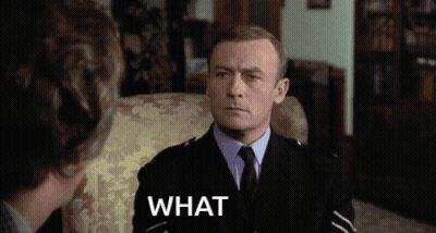 A Man In A Suit And Tie Is Sitting In A Chair With The Word What Written Below Him GIF