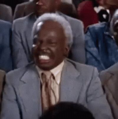 A Man In A Suit And Tie Is Sitting In A Crowd Of People And Laughing . GIF