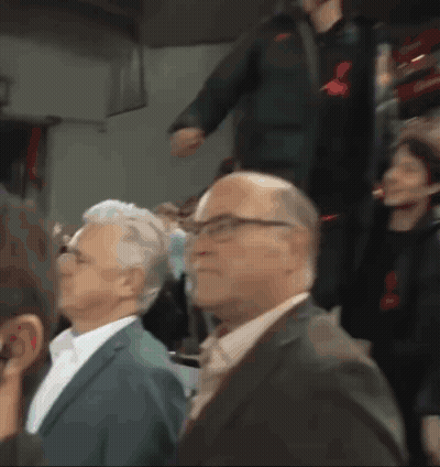 A Man In A Suit And Tie Is Sitting In A Crowd Of People GIF