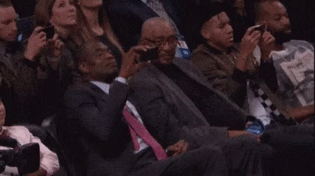 A Man In A Suit And Tie Is Sitting In A Crowd Of People Watching A Basketball Game . GIF