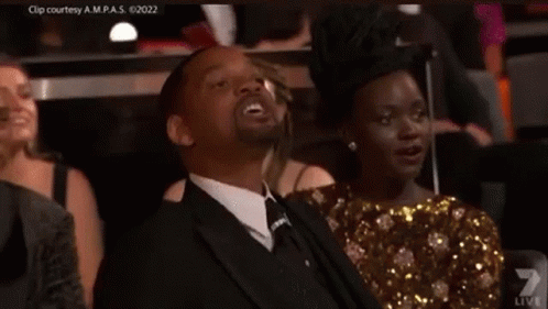 A Man In A Suit And Tie Is Sitting In A Theatre With A Woman . GIF