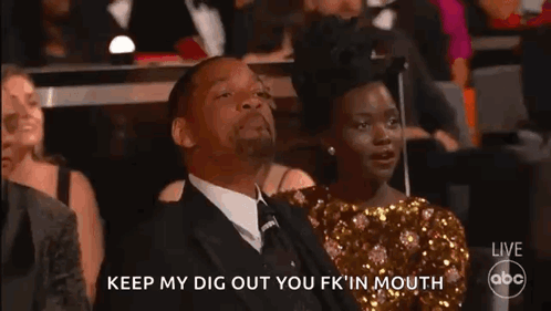 A Man In A Suit And Tie Is Sitting In A Theatre With A Woman In A Gold Dress Behind Him . GIF