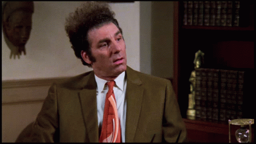 A Man In A Suit And Tie Is Sitting In Front Of A Bookcase GIF