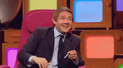 A Man In A Suit And Tie Is Sitting In Front Of A Bunch Of Televisions And Smoking A Cigarette . GIF