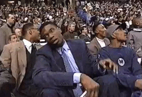 A Man In A Suit And Tie Is Sitting In Front Of A Crowd At A Basketball Game . GIF