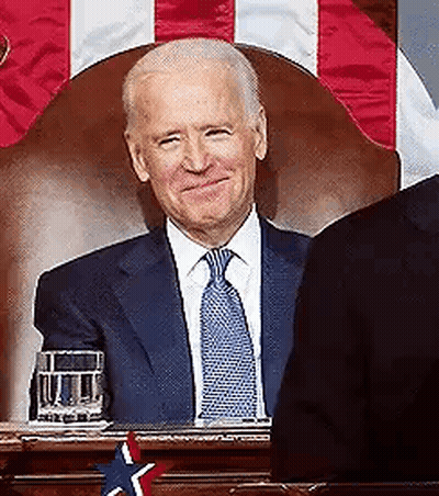 A Man In A Suit And Tie Is Sitting In Front Of A Flag . GIF