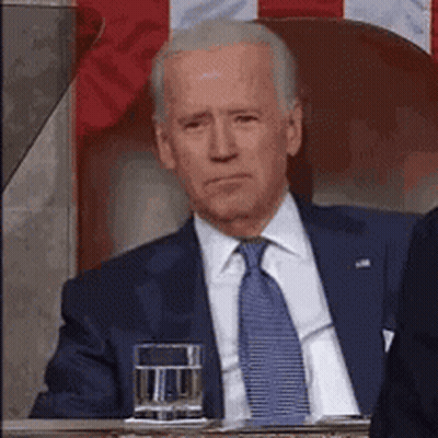 A Man In A Suit And Tie Is Sitting In Front Of A Flag With A Glass Of Water . GIF