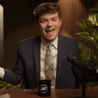 A Man In A Suit And Tie Is Sitting In Front Of A Microphone And A Mug That Says Af GIF
