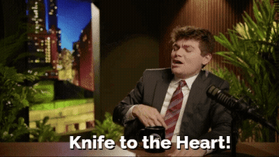 A Man In A Suit And Tie Is Sitting In Front Of A Microphone And Says Knife To The Heart GIF