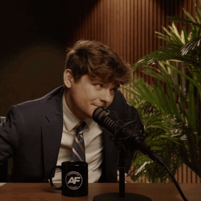 A Man In A Suit And Tie Is Sitting In Front Of A Microphone With A Black Mug That Says Af On It GIF