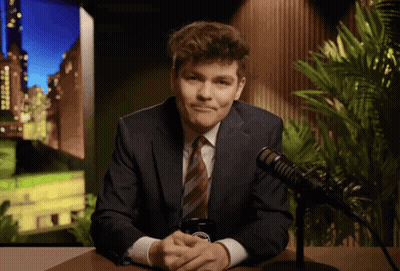 A Man In A Suit And Tie Is Sitting In Front Of A Microphone With A Cell Phone In His Hand GIF