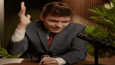 A Man In A Suit And Tie Is Sitting In Front Of A Microphone With A Coffee Mug On The Table GIF