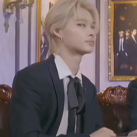 A Man In A Suit And Tie Is Sitting In Front Of A Picture On The Wall . GIF