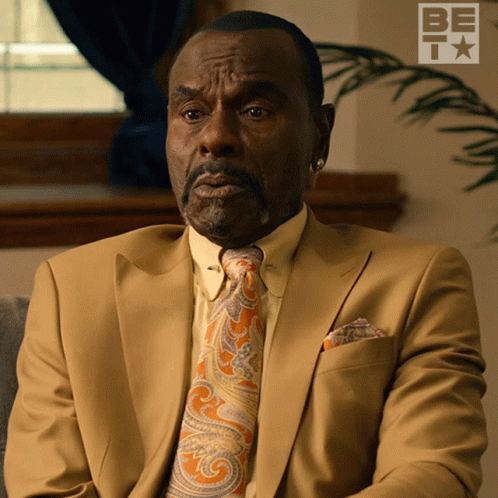 A Man In A Suit And Tie Is Sitting In Front Of A Sign That Says Be GIF