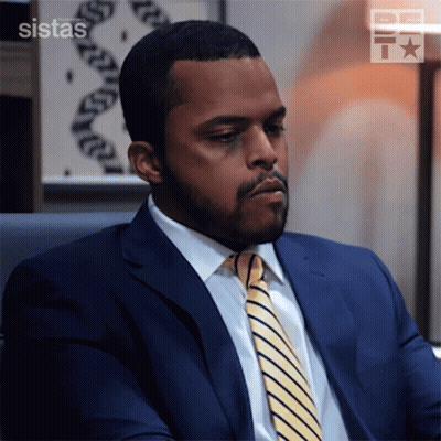 A Man In A Suit And Tie Is Sitting In Front Of A Sign That Says ' Sistas ' GIF