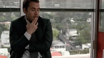 A Man In A Suit And Tie Is Sitting In Front Of A Window And Covering His Mouth With His Hand . GIF