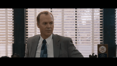 A Man In A Suit And Tie Is Sitting In Front Of A Window With Blinds GIF